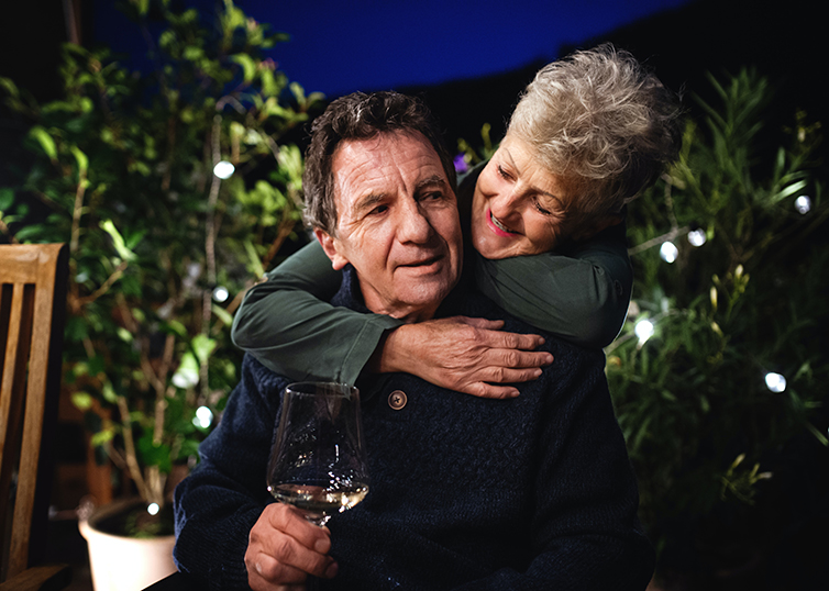 an older couple with the man sitting with a wine glass and the woman hugging from behind