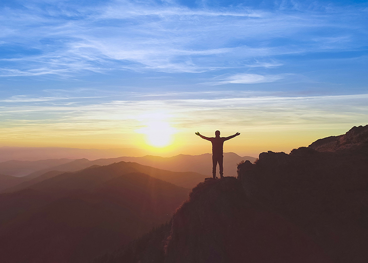 person on a cliff with arms outstretched with sunset in background